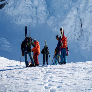 Backcountry skiers preparing below a sunlit snow cornice