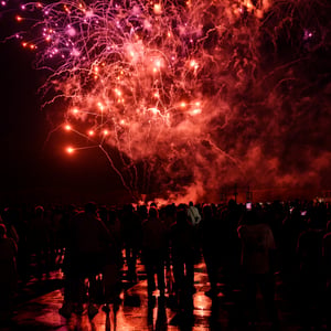 Crowd watching red fireworks over a wet plaza