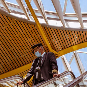 Pilot in face mask riding airport escalator with suitcase