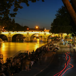 Crowds on the Seine riverbank beneath a lit stone bridge